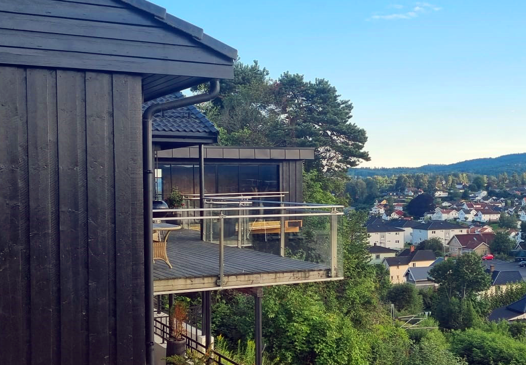 En premier plan, il y a la maison principale et sa terrasse en bois. En arrière plan, on voit l'extension s'avancée dans le vide au dessus de la montagne.